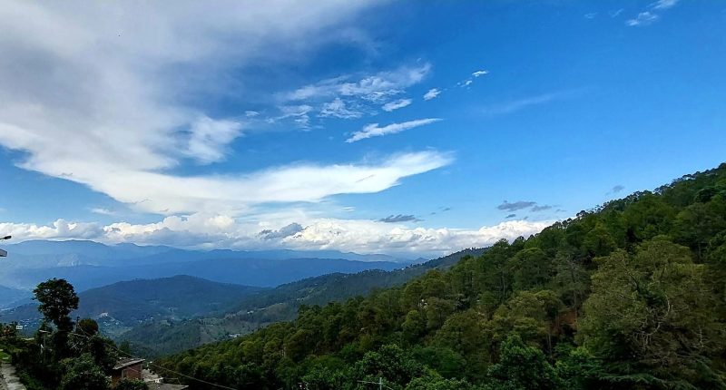 Mountain valley under a blue sky with scattered clouds; forested hillside on the right and small buildings at the bottom left.