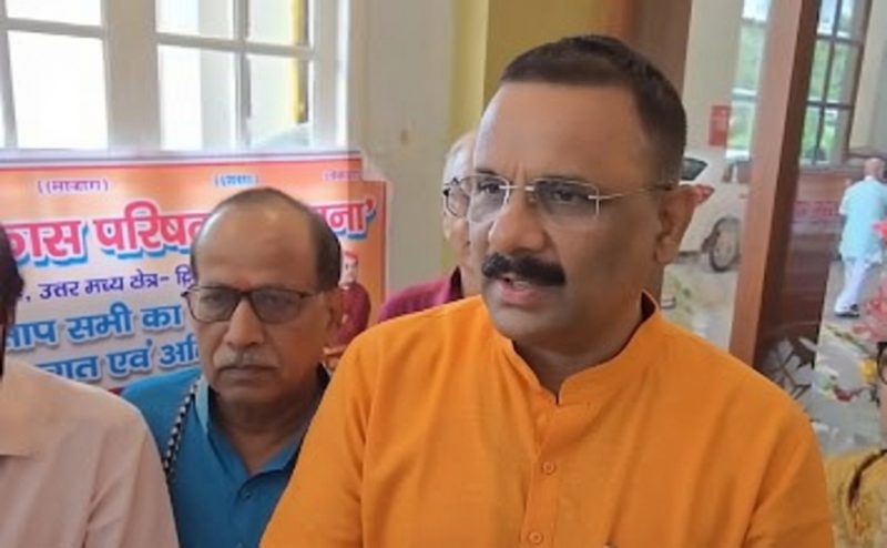 Man in an orange kurta wearing glasses speaks to an audience indoors, with a poster and a man in a blue shirt in the background.