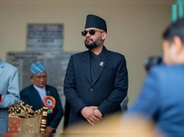 Bearded man in a black suit and Nepali topi wearing sunglasses at a formal event, with ceremonial objects and other attendees in the background.