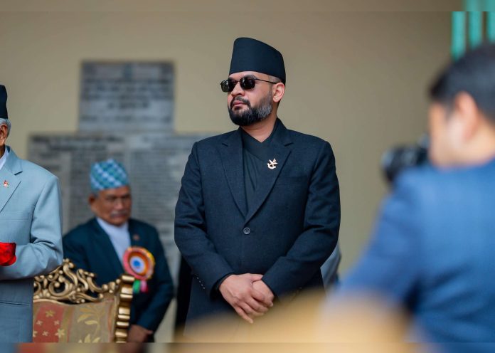 Bearded man in a black suit and Nepali topi wearing sunglasses at a formal event, with ceremonial objects and other attendees in the background.
