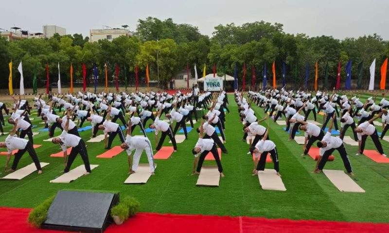 crpf s - bolebharat Large outdoor yoga event with rows of participants on colored mats performing a side bend on a green lawn, colorful flags in the background.