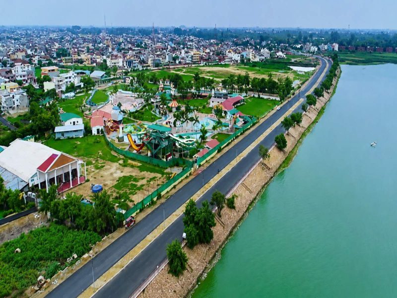 gorakhpur 2 - bolebharat Aerial view of a riverside park with playgrounds, palm trees, and green lawns, beside a curved road along the water.