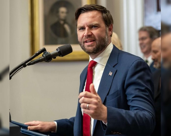 Man in a navy suit and red tie speaks at a podium with a microphone indoors.