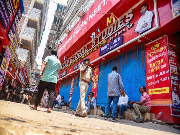Busy Indian street with closed blue storefront shutters and bright red shop signage; a police officer talks on his mobile while pedestrians stroll and sit nearby.