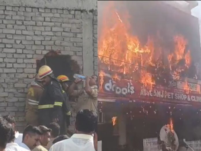 Firefighters in protective gear battle a building fire as onlookers watch nearby by a brick wall.