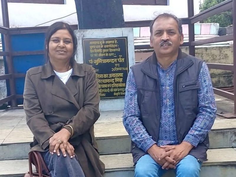 Two adults sit side by side on outdoor concrete steps, with a plaque behind them in a courtyard setting.