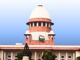 India’s national flag flies in front of a red-brick domed government building (parliament/Capitol-style) against a blue sky.