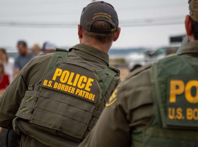 Two U.S. Border Patrol officers in tactical vests, seen from behind at a border event.