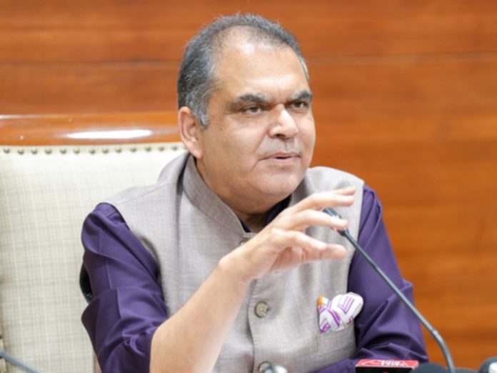Man in a beige waistcoat and purple shirt speaks into a microphone at a press conference, with a wooden paneled background.