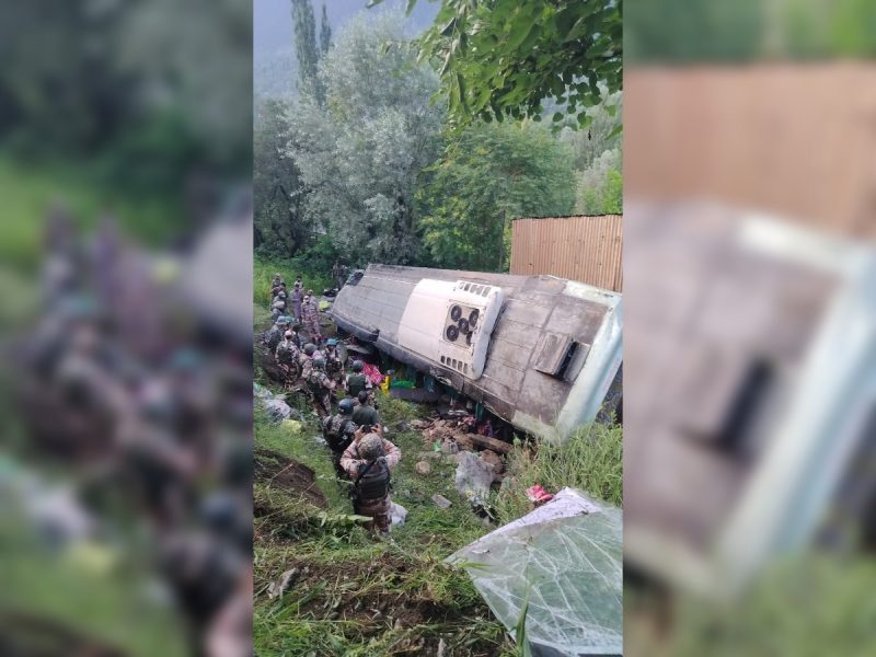 Overturned bus on its side in a grassy, hilly area with soldiers and bystanders nearby.