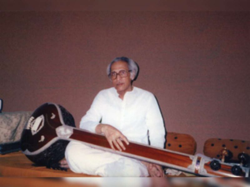Older man in a white kurta sits cross-legged on the floor holding a sitar-like string instrument.