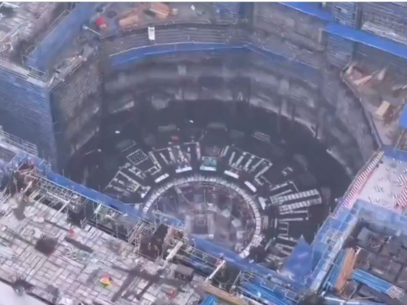 Aerial view of a deep circular excavation with concentric platforms and blue safety barriers under construction, viewed from above.