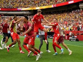 Soccer players in red jerseys celebrate on the field as a teammate jumps with others around him.
