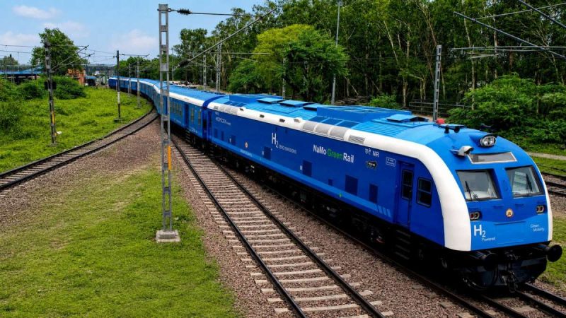 Blue hydrogen-powered passenger train curving along tracks through a green landscape on an overhead-line route.