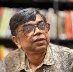 Close-up of a middle-aged man with gray hair and large glasses, in a library backdrop with blurred bookshelves.