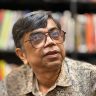 Close-up of a middle-aged man with gray hair and large glasses, in a library backdrop with blurred bookshelves.
