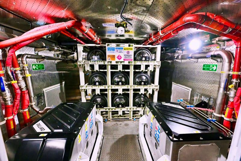Interior of a maintenance compartment with red insulated pipes, metal walls, and two large front-loading machines in the foreground; a rack of circular components sits at the far end under various warning signs.