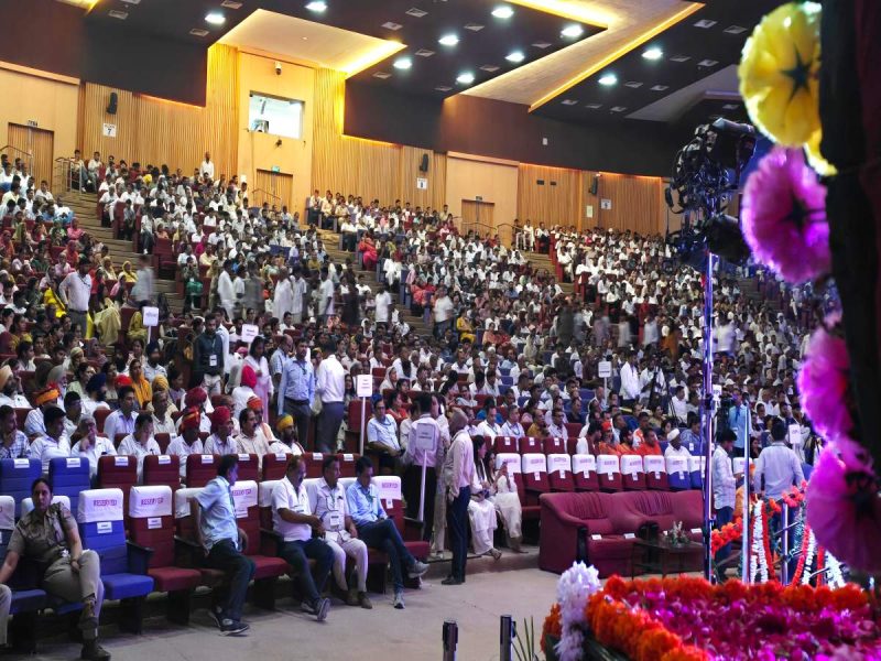 Large auditorium filled with a diverse crowd seated in rows, facing a stage on the right with colorful flower decorations.