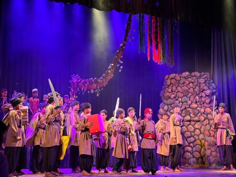 Group of children in traditional robes performing on a theater stage, holding swords and props with a colorful backdrop and stone set piece.