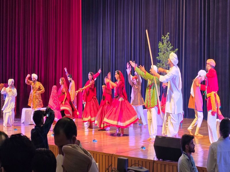 Group of performers in bright traditional Indian attire dancing on a stage with red and blue curtains behind them.