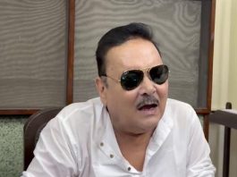 Man wearing a white shirt and dark sunglasses, talking or mid-speech indoors.