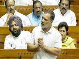 Man in a white polo stands at the front of a parliamentary chamber, hands pressed together while colleagues wearing headsets sit behind him.