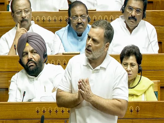 Man in a white polo stands at the front of a parliamentary chamber, hands pressed together while colleagues wearing headsets sit behind him.