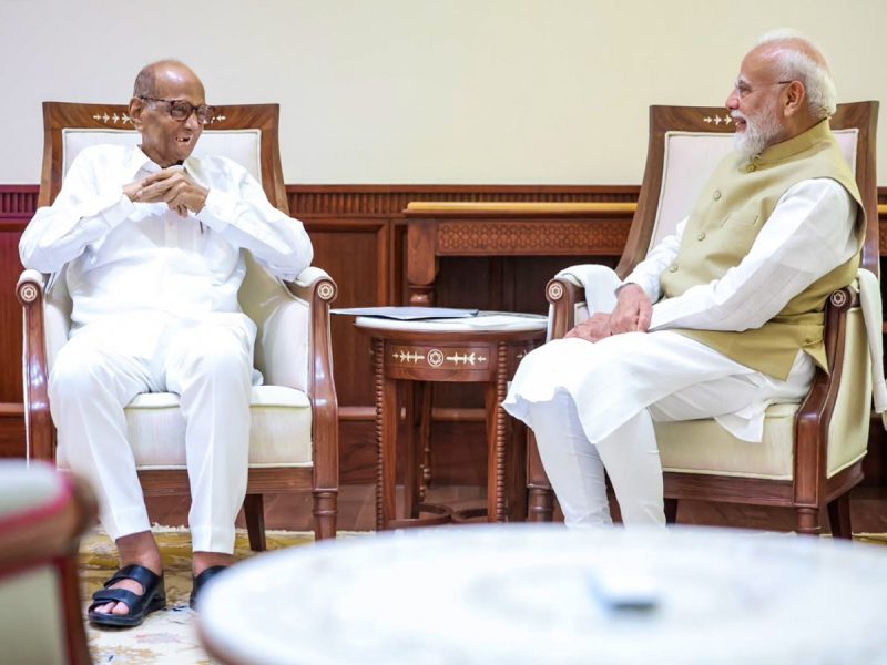 Two men sit in ornate chairs facing each other, smiling and talking in a formal room with a wooden table between them.