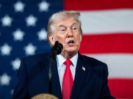 A man in a dark suit and red tie speaks at a podium with an American flag in the background, giving a speech.