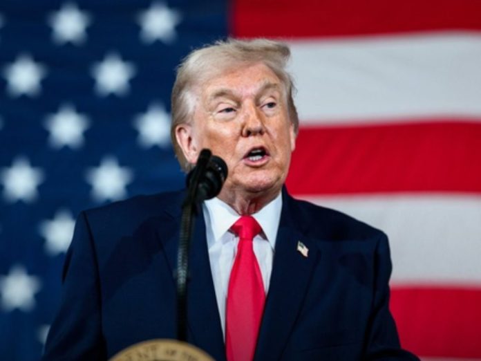 A man in a dark suit and red tie speaks at a podium with an American flag in the background, giving a speech.