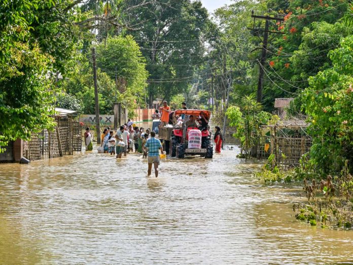 Flooded village street with people wading in knee- to waist-deep water and a tractor carrying passengers in the background.