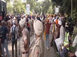 Large street protest with police officers and a crowd of Sikhs wearing orange turbans, some holding signs while media cameras film.