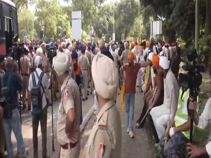 Large street protest with police officers and a crowd of Sikhs wearing orange turbans, some holding signs while media cameras film.