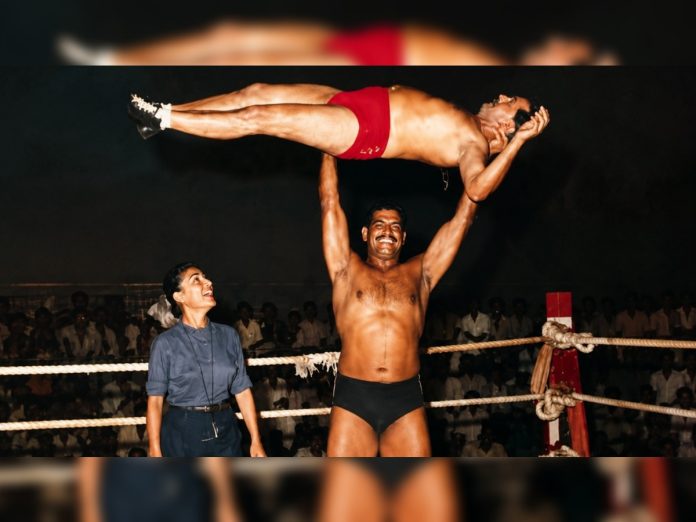 Muscular wrestler in black trunks lifts a partner in red trunks overhead in a ring while a woman spectator watches nearby.