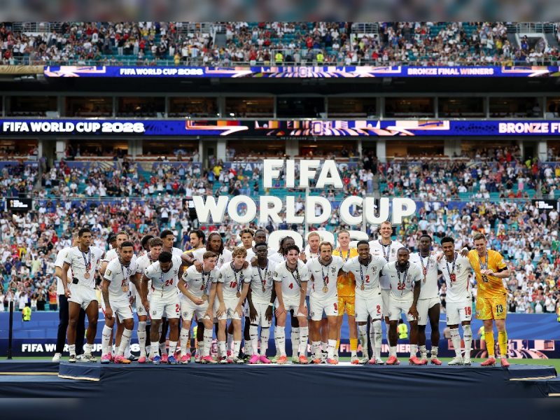 image 3 - bolebharat National soccer team players in white uniforms pose on a podium with medals at a FIFA World Cup 2026 event, large 'FIFA WORLD CUP' sign behind them, stadium crowd in the background.
