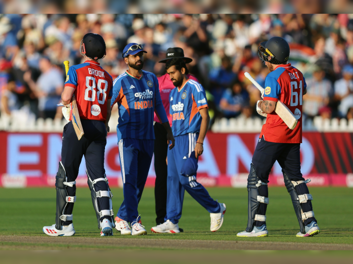 Cricket match scene with India players in blue uniforms and opposing team in red, batsmen with pads on the field and a crowded stadium in the background.