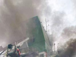 A large ship or ship-side structure on fire, with thick smoke and tilted green hull amid emergency scenes on deck equipment.