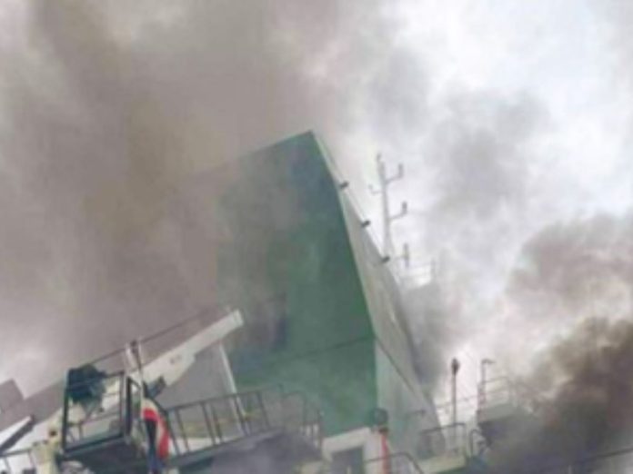 A large ship or ship-side structure on fire, with thick smoke and tilted green hull amid emergency scenes on deck equipment.