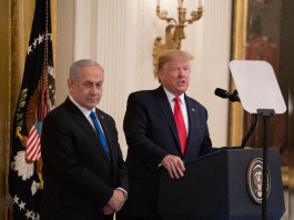 Two men in dark suits stand at a podium with the presidential seal; one speaks into a microphone in a grand room with flags in the background.