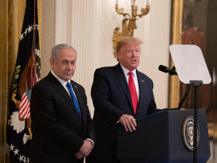 Two men in dark suits stand at a podium with the presidential seal; one speaks into a microphone in a grand room with flags in the background.