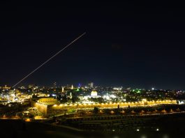 Nighttime cityscape with warm lights along ancient walls, a domed building in the center, and a long diagonal light trail across the dark sky.
