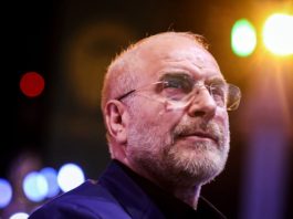 Man with glasses and gray beard looking upward on stage with colorful stage lights in the background, nighttime concert vibe.