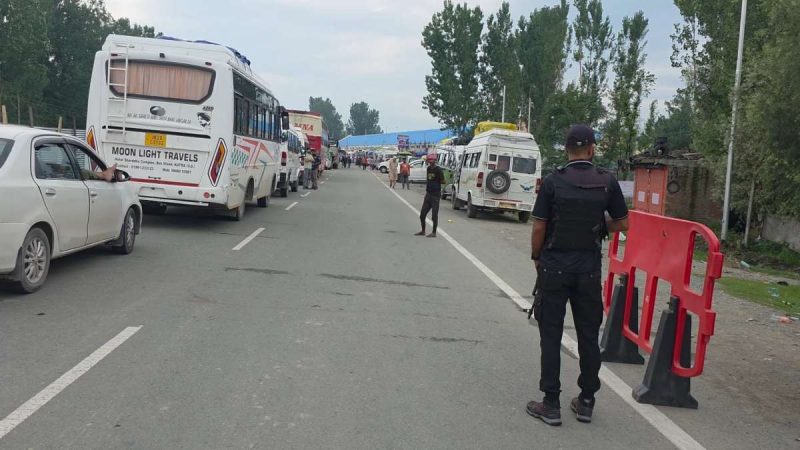 Security checkpoint on a road with parked buses and cars; a uniformed officer stands near a red barrier in the foreground.