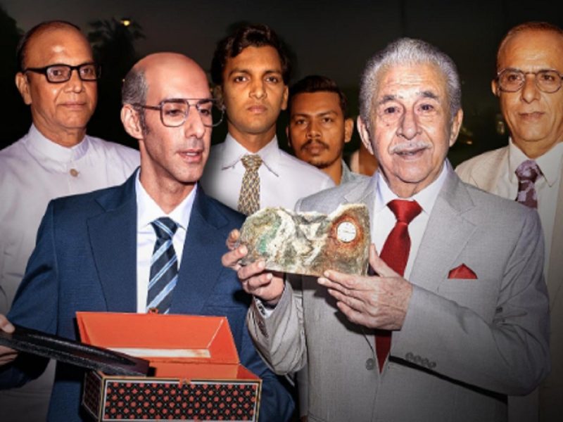Group of men in suits posing at an awards event; two men in front hold a stone trophy and a gift box.