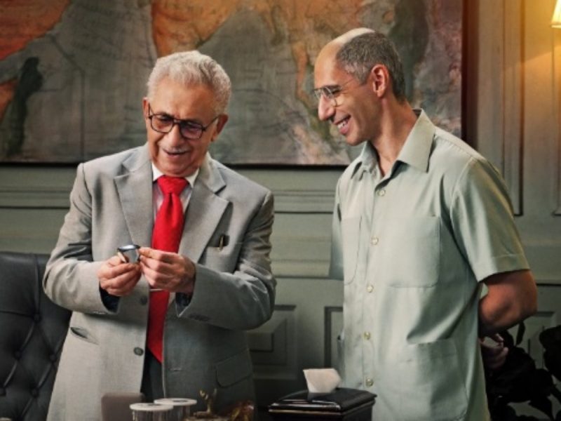 Two men in an office: an older man in a gray suit and red tie shows a small device to a younger man in a light green shirt, smiling as they look at it on the table.