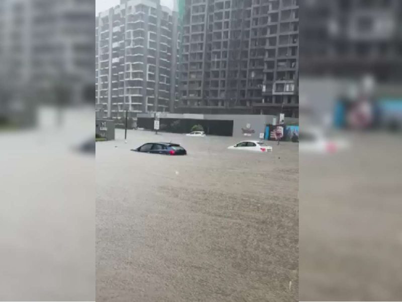 Flooded city street with partially submerged cars and high-rise buildings under construction in the background, gray rainy scene.