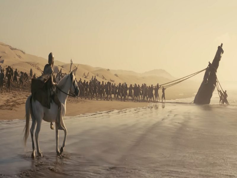 Rider on a white horse watches as a long line of people on the beach drag a tall ship or mast toward the sea at sunset. Desert dunes rise in the background.