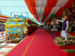 A man in traditional Indian attire waves a green flag beside a decorated train during an inauguration ceremony under red and white drapes.