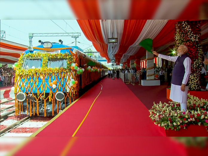 A man in traditional Indian attire waves a green flag beside a decorated train during an inauguration ceremony under red and white drapes.