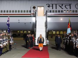 Dignitary walks a red carpet away from a large aircraft with 'Bharat' on the fuselage, flanked by soldiers at attention.